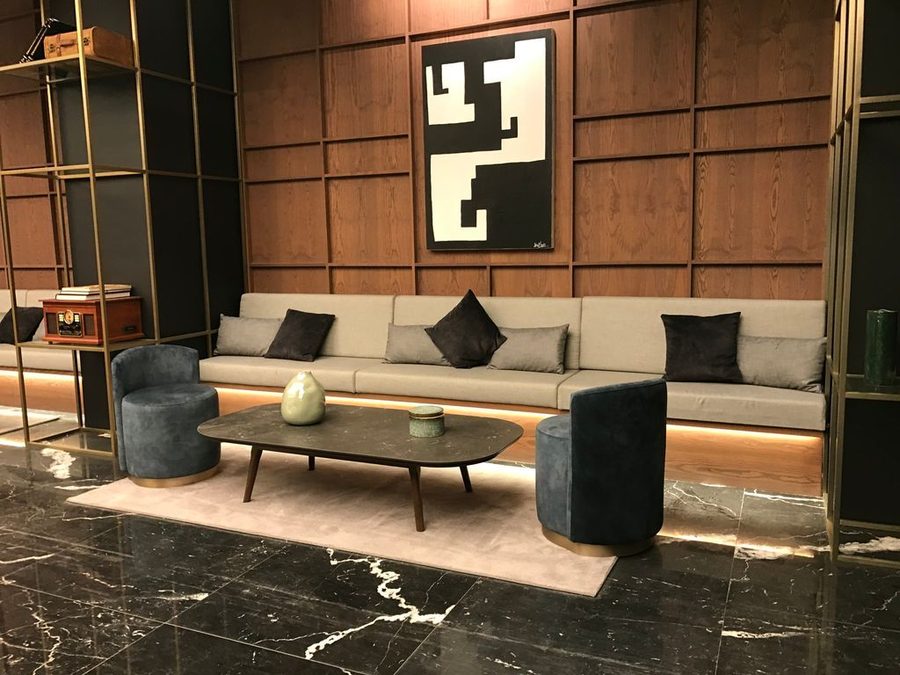 Lounge de hotel revestido a madeira: banqueta comprida em tom claro, maples giratórios e poufs sobre tapete.