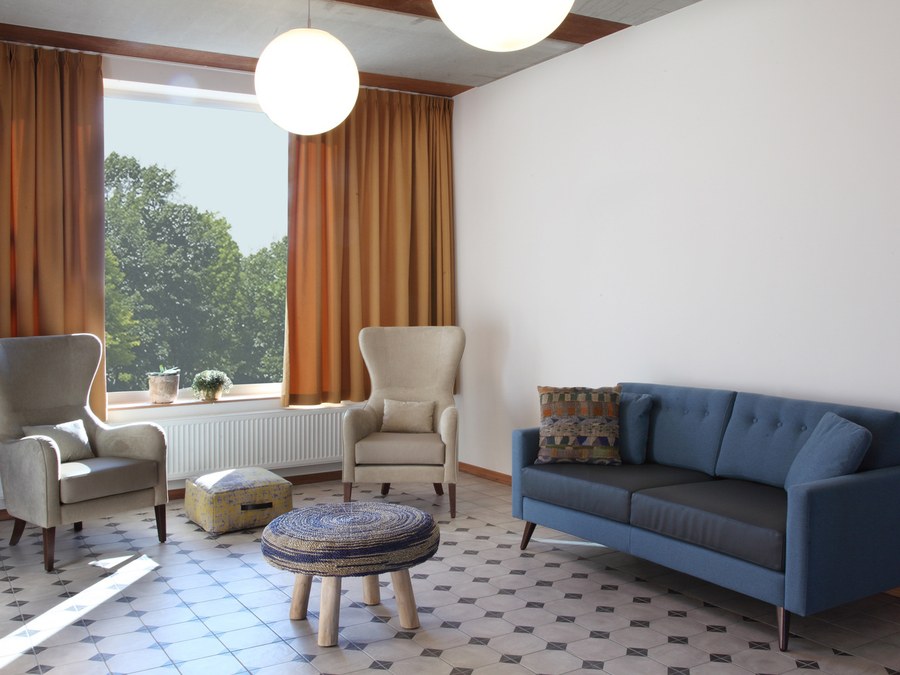 Sala de estar de um lar: dois maples de orelhas em bege claro diante de uma janela com cortinados âmbar, frente a um sofá azul, um pouf amarelo e um banco entrançado, sobre um mosaico com padrão.