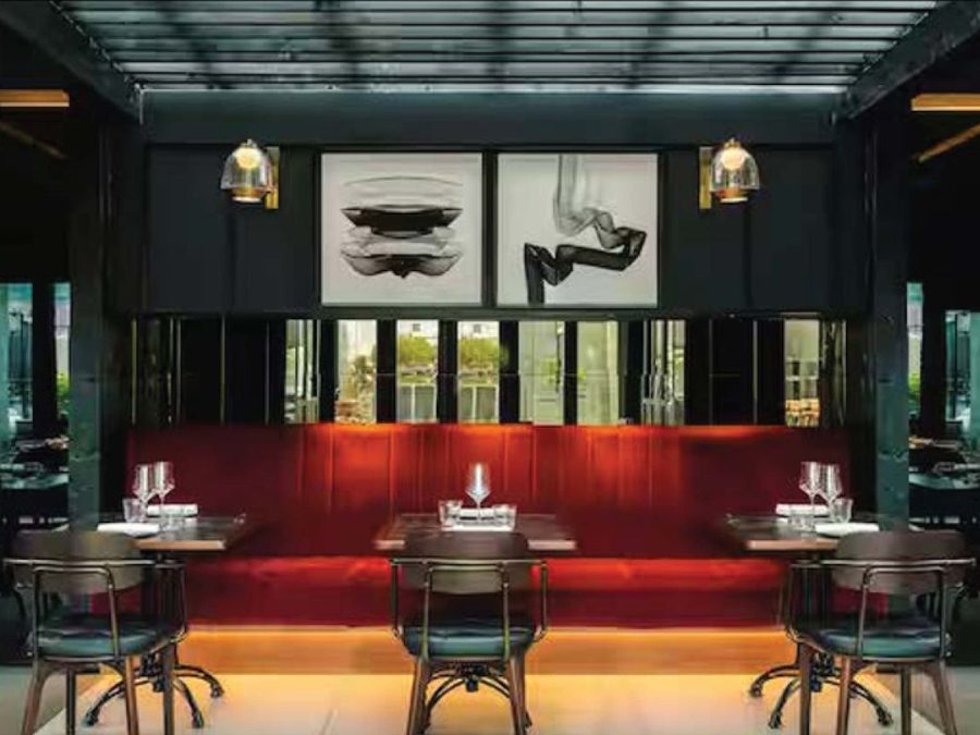 Sala de restaurante: banqueta comprida em veludo vermelho, mesas e cadeiras em metal diante de uma envidraçada preta.
